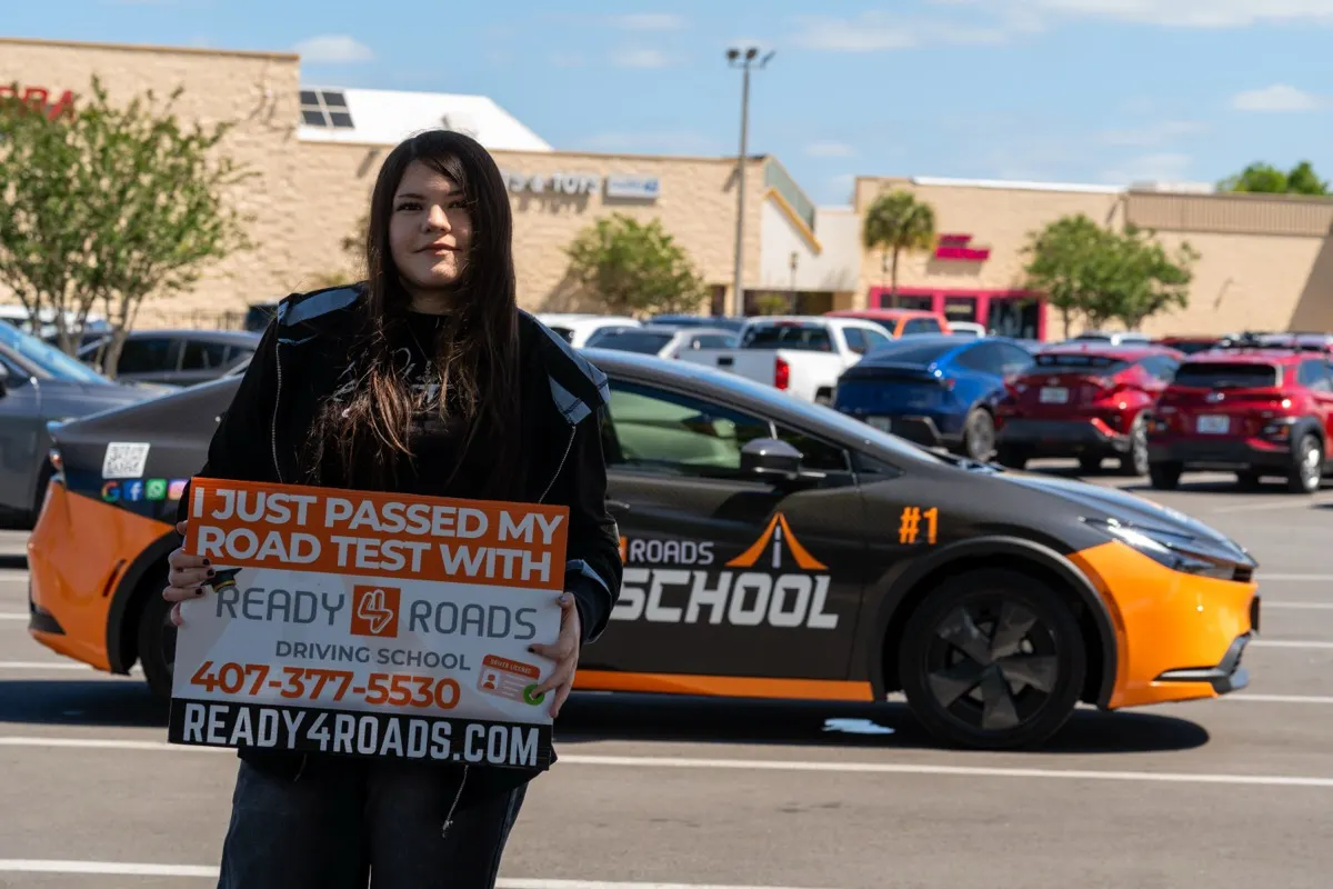 DMV road test prep in Kissimmee, FL · Ready 4 Roads student holding an “I just passed my road test” sign next to a training car